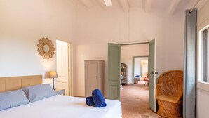 A bedroom with a bed, a nightstand, a lamp, a wardrobe, and a wicker chair. - Costabravaforrent Tallada (La Tallada d'Empordà)