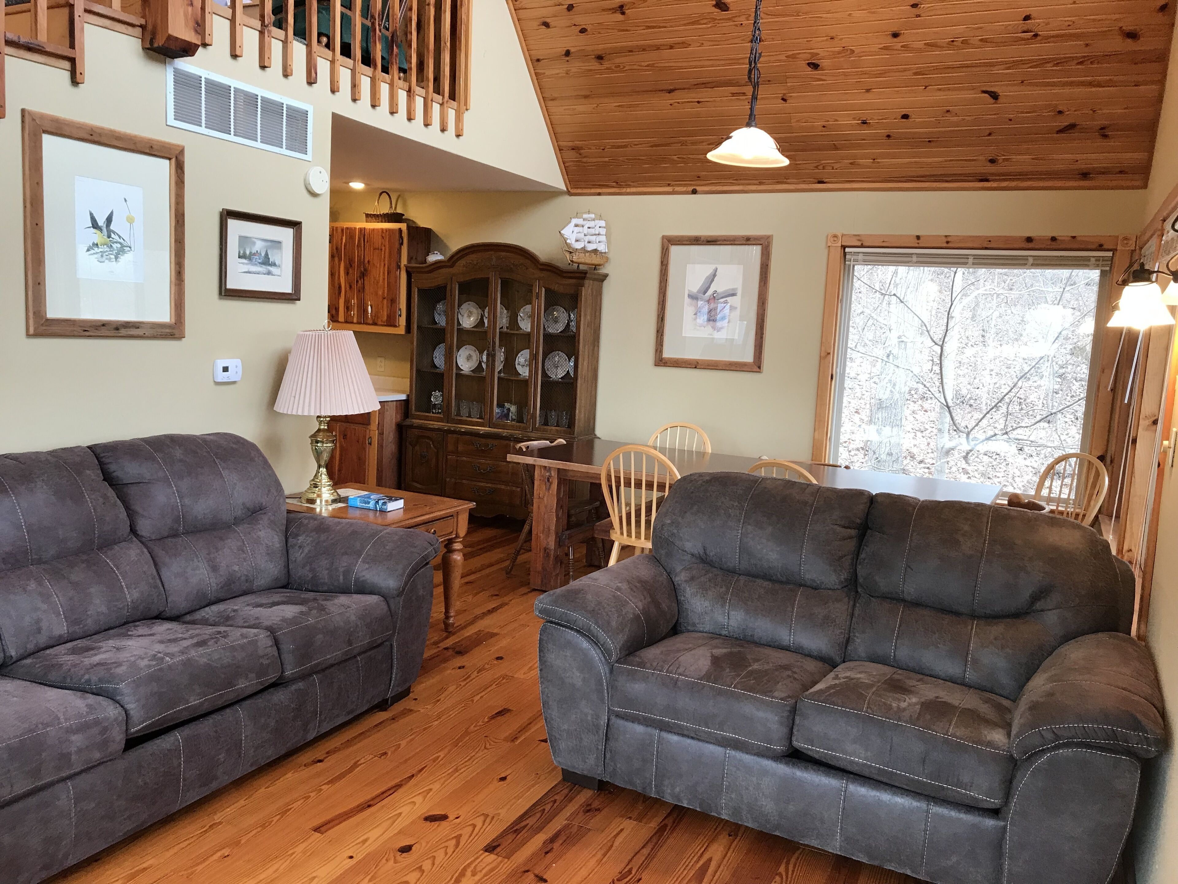 A cozy living room with two dark sofas, wooden flooring, a dining table with chairs, a wooden cabinet with glass doors, and a large window with a view of trees.
