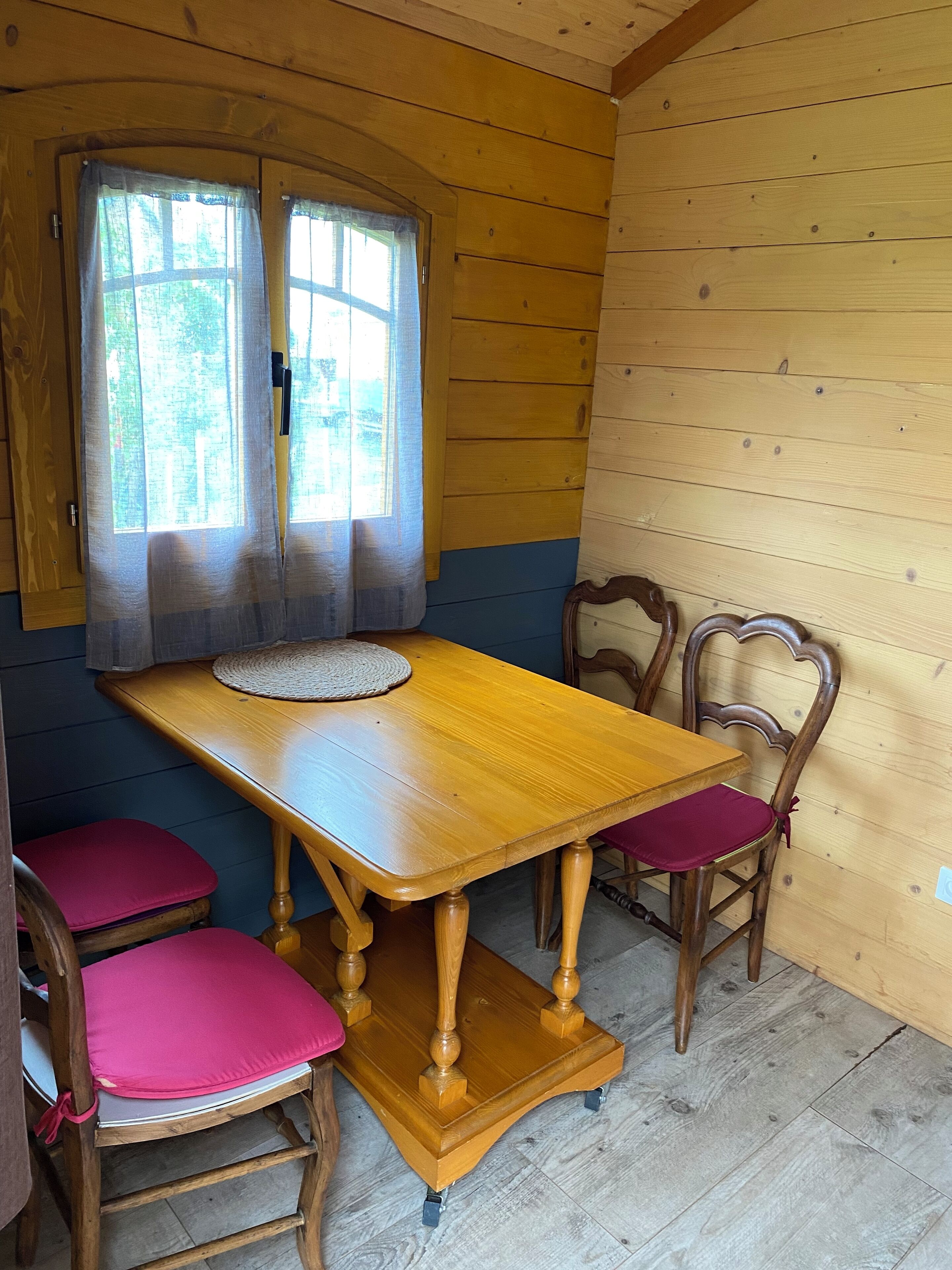 A wooden dining room with a table and chairs, a window with curtains, and a view of greenery outside.