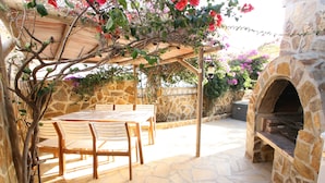 Um pátio ao ar livre com piso de pedra, pérgola de madeira, forno de pedra e uma mesa com cadeiras.