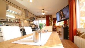 Uma sala de jantar com uma mesa de madeira, cadeiras, uma televisão e uma cozinha ao fundo.