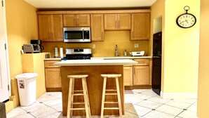 Una cocina con armarios de madera, un microondas de acero inoxidable, un refrigerador negro y un reloj en la pared.