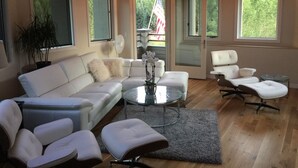 Una sala de estar moderna con un sofá seccional blanco, una mesa de centro de vidrio y una alfombra gris. Hay dos sillones blancos y una pequeña mesa redonda.
