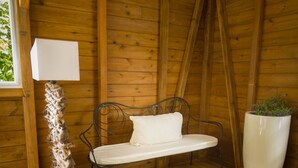 A cozy wooden cabin interior with a metal bench, a white cushion, a large potted plant, and a small table with a vase and a teapot. - Locastudio "Pomme Kanel (Sainte-Anne)