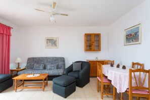 A living room with a sofa, armchair, ottoman, wooden coffee table, dining table with chairs, a cabinet, and a ceiling fan.