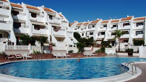 A swimming pool surrounded by white buildings with red-tiled roofs, a clear blue sky, and a brick pathway. - Sunny 2-Bedrooms With Large Terrace. WiFi (Arona)