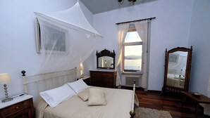 A bedroom with a bed, bedside tables, a nightstand, a window with curtains, a mirror, and a view of the sea.