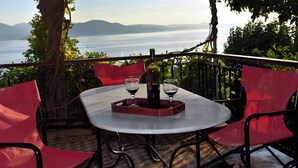 A round table with a bottle of wine and two glasses on a tray, surrounded by red chairs, overlooking a body of water with mountains in the distance.