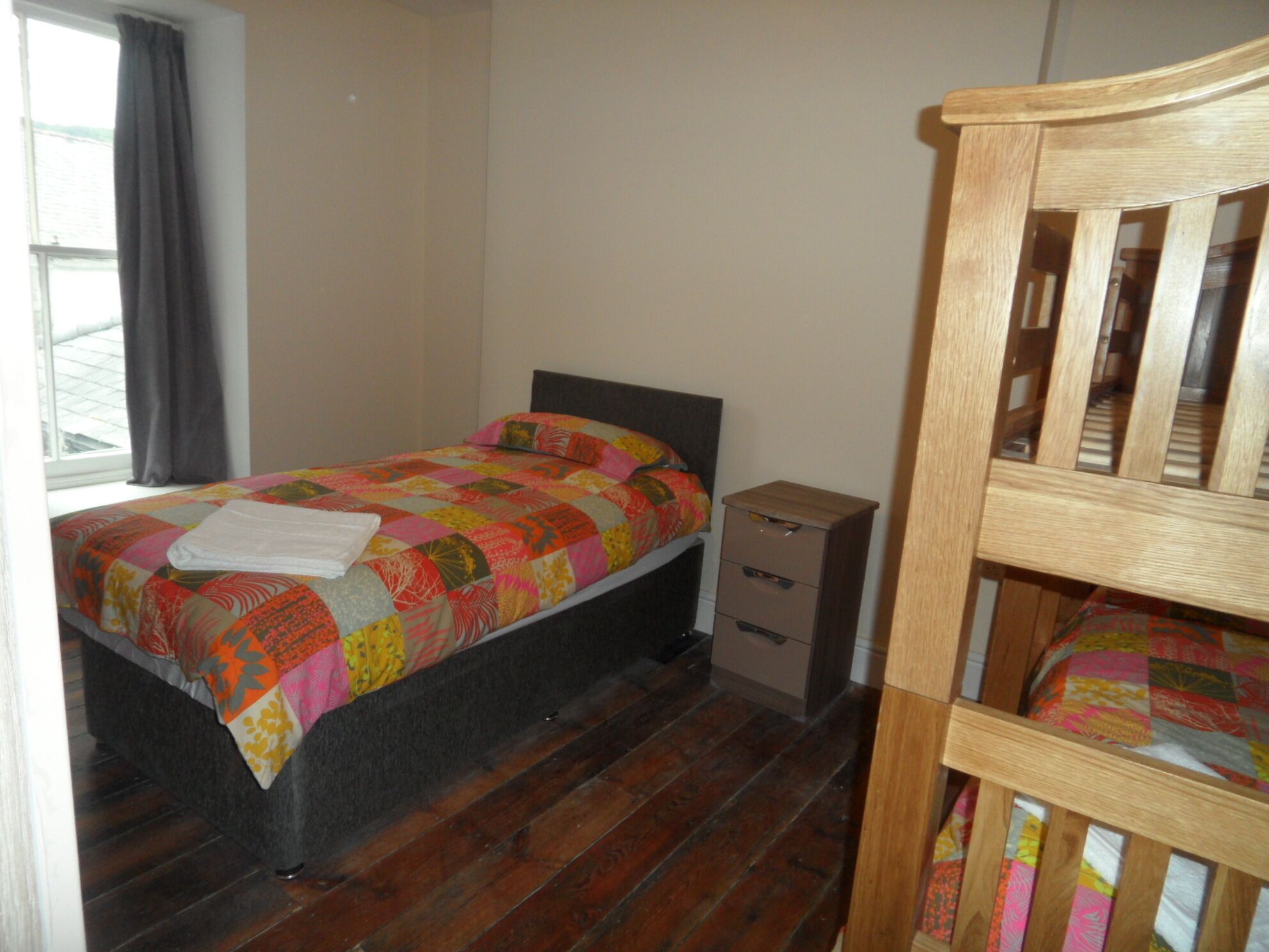 Un dormitorio con cama, mesita de noche, silla de madera y ventana con cortinas.