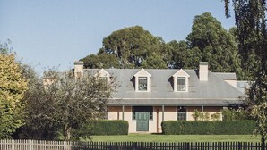 Une maison avec un étang au premier plan, entourée d’herbes hautes et d’arbres.