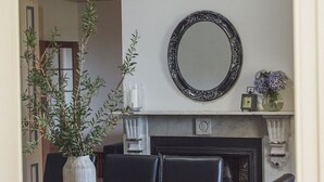 Un coin repas avec une table en bois, des chaises noires, un vase blanc orné de feuillage vert, un miroir décoratif et une cheminée sur laquelle sont posés une horloge et des fleurs.