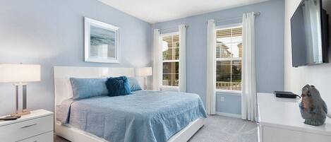A bedroom with a bed, a nightstand, a dresser, a flat-screen TV, and a window with curtains.