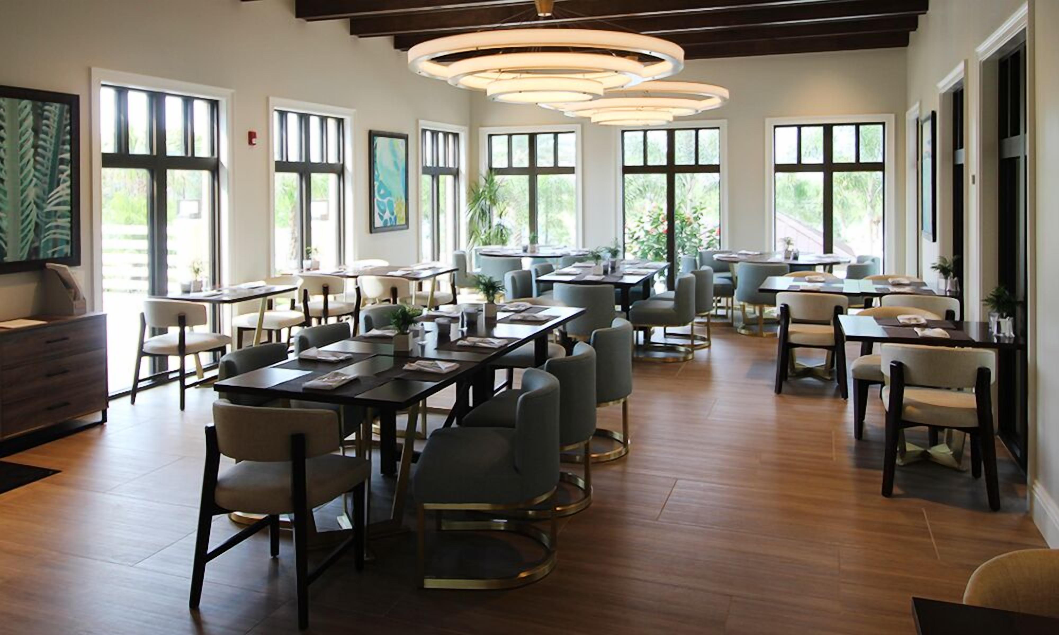 A spacious dining area with multiple tables and chairs, large windows allowing natural light, and a modern ceiling light fixture.