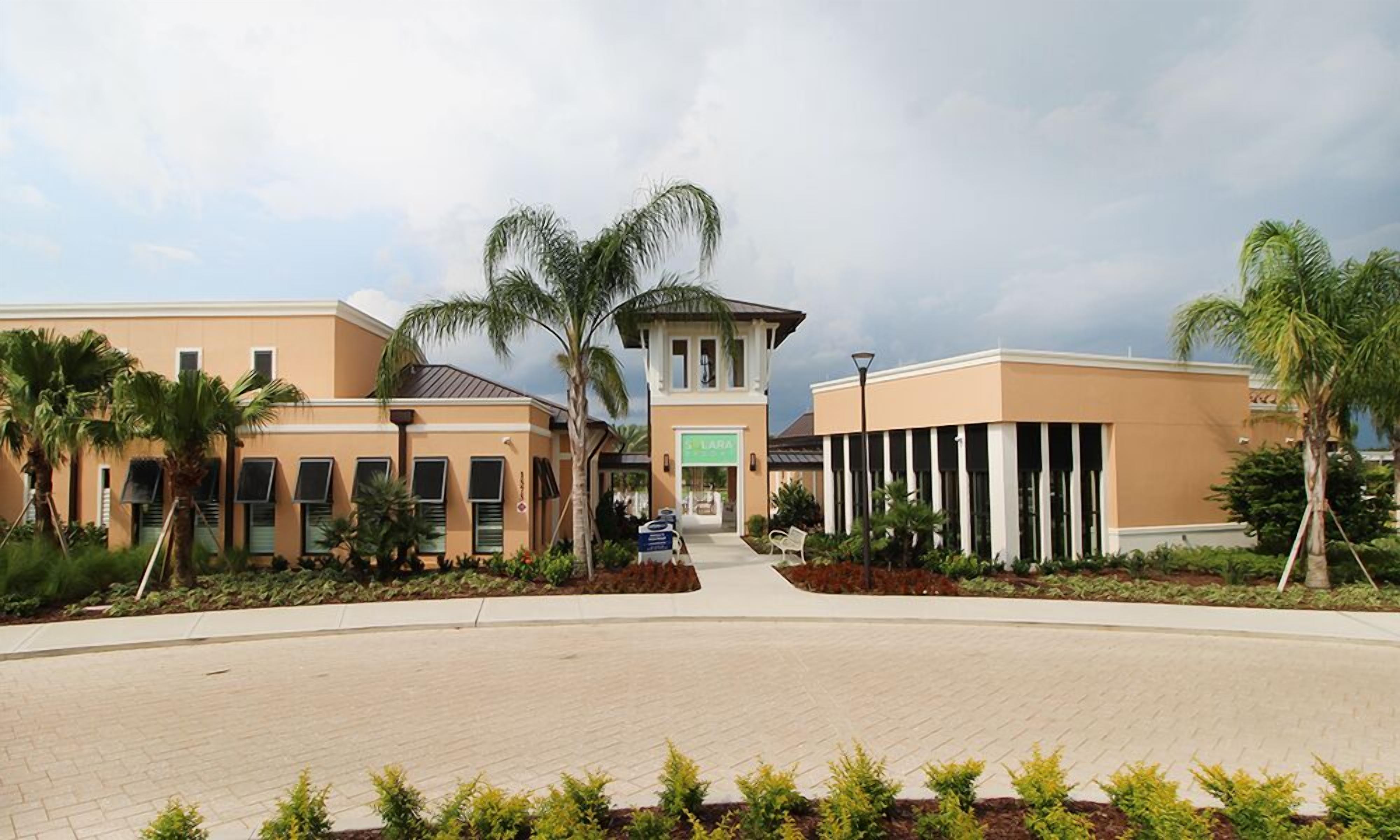 A hotel with a central tower, two wings, and a paved courtyard with palm trees.