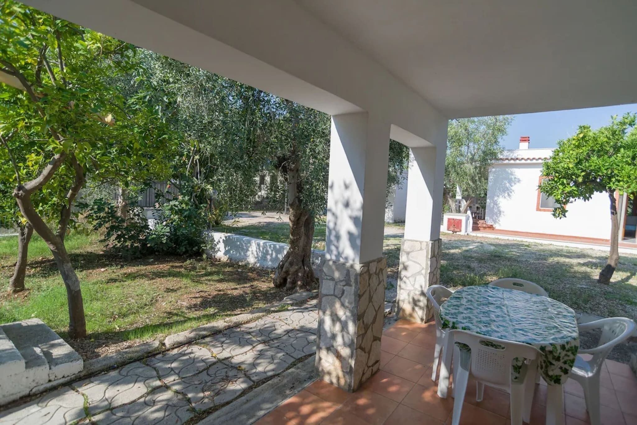 A covered outdoor area with a stone-tiled patio, a round table with chairs, and surrounding greenery.
