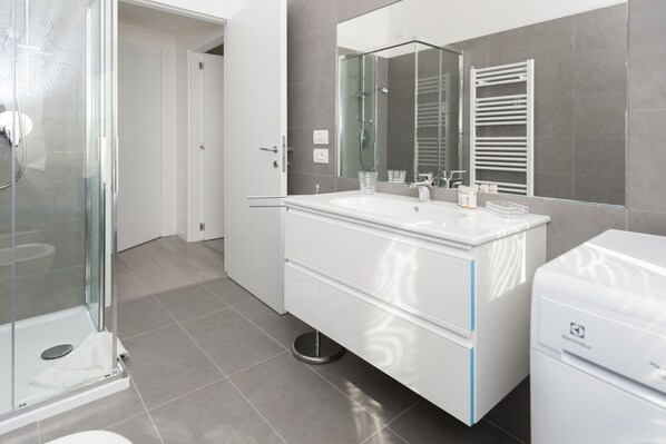 A modern bathroom with a glass shower enclosure, a white vanity with a large mirror, and a white washing machine.