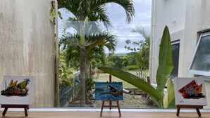 Une pièce dotée d’une grande fenêtre donnant sur un jardin planté de palmiers, et où est exposé un tableau sur un chevalet.