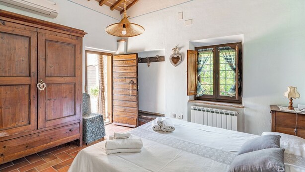 A bedroom with a wooden wardrobe, a bed with white linens, a radiator, and a window with curtains. - House in the Cortona countryside. Romantic Little House (Fratticciola)