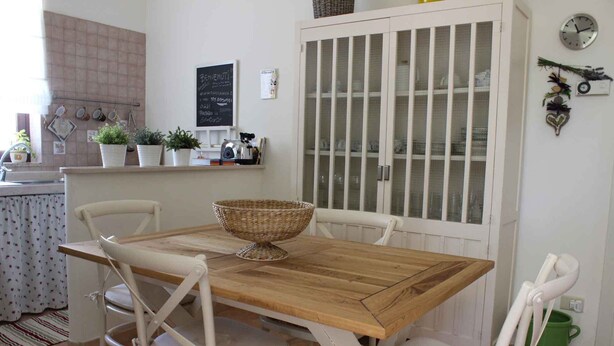 A dining area with a wooden table and white chairs, a cabinet with glass doors, and a kitchen area with hanging pots and a chalkboard menu. - Vacation House Fiordilavanda (Martina Franca)