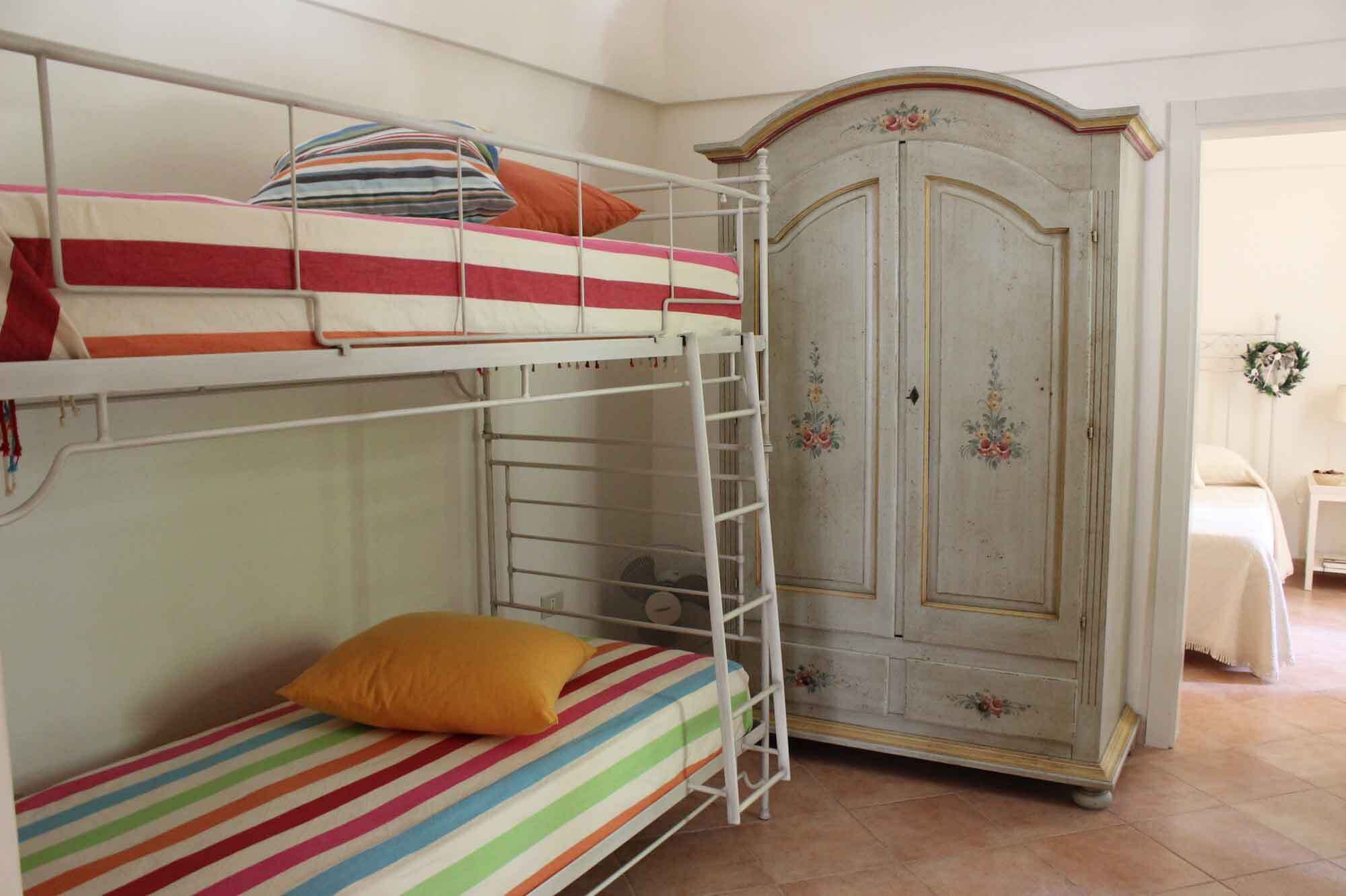 Ein Etagenbett mit einer Stehle, ein Einzelbett mit einer farbenfrohen, gestreiften Bettdecke und ein antiker Kleiderschrank mit floralen Verzierungen.