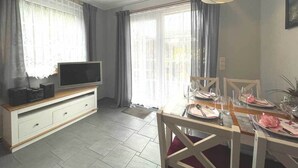 Un coin repas avec une table en bois dressée pour deux personnes, un téléviseur posé sur un meuble blanc et une grande fenêtre agrémentée de rideaux.