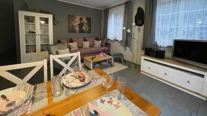Un salon comprenant une table à manger dressée pour deux personnes, un canapé, un meuble avec des portes en verre, un téléviseur et une fenêtre avec des rideaux.