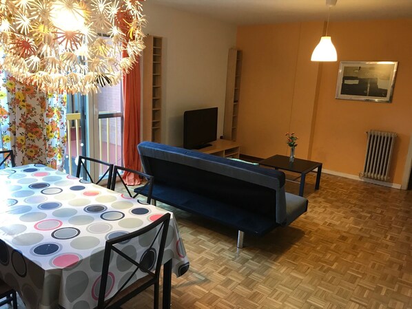 Un salon avec un canapé bleu, une table à manger recouverte d’une nappe à pois, un meuble pour la télévision et un radiateur.