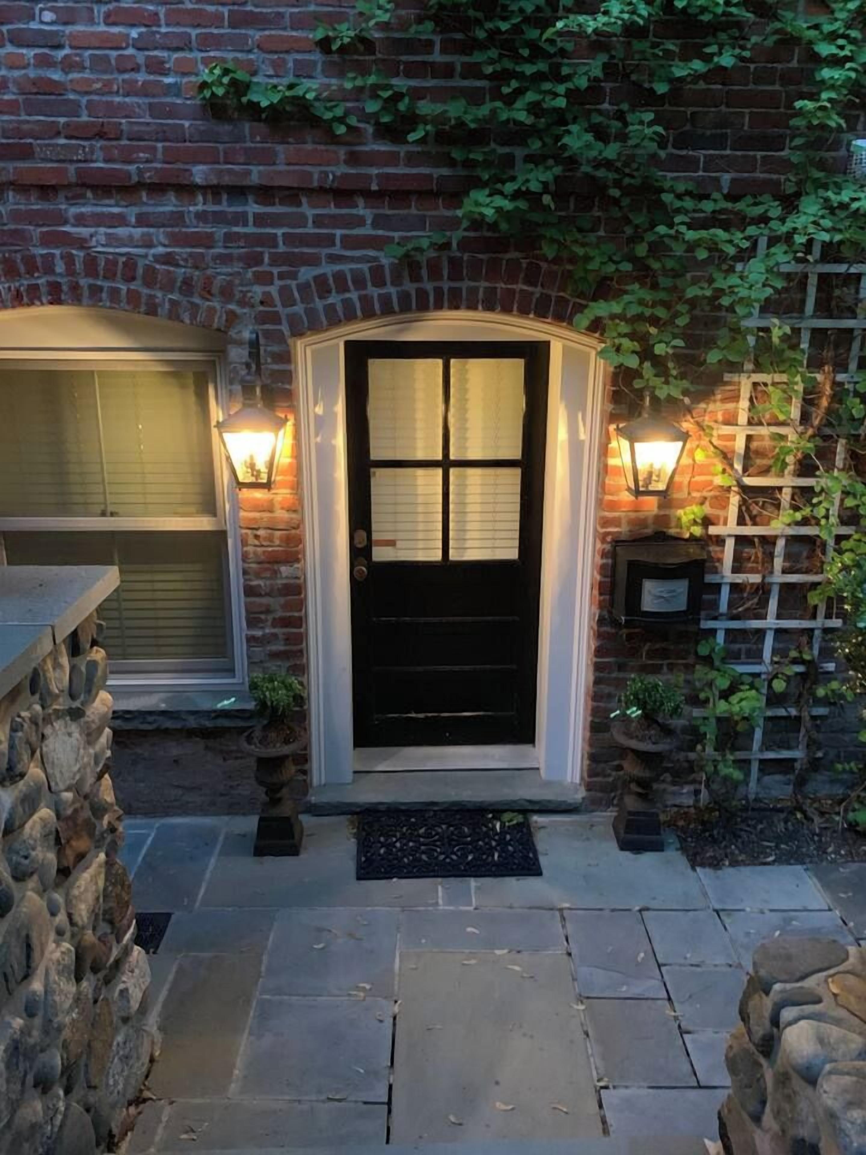 A brick house with a black front door, two wall-mounted lights, and a stone wall on the left.