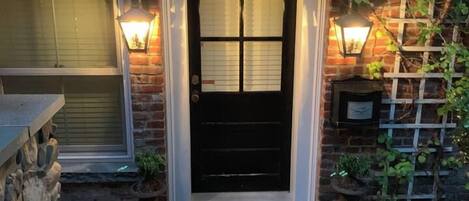 A brick house with a black front door, two wall-mounted lights, and a stone wall on the left.
