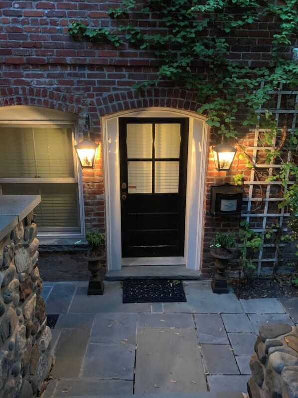 A brick house with a black front door, two wall-mounted lights, and a stone wall on the left.