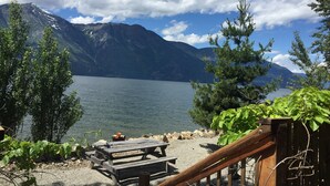 A lakeside view with a wooden deck, picnic table, and benches overlooking a calm lake and mountains.