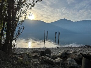 Blick auf einen See mit Bergen im Hintergrund, hölzernen Pfählen im Wasser und einer felsigen Uferlinie.