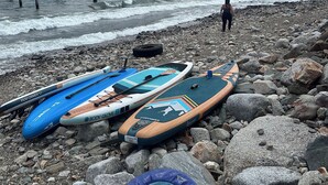 A rocky beach with kayaks and a paddleboard, mountains in the background, and a person standing in the distance.