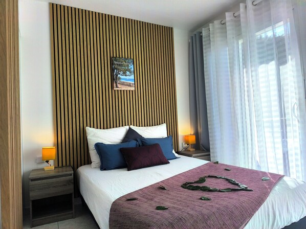 A neatly made bed with a white comforter and multiple pillows, a wooden slat wall panel, a small bedside table with a lamp, and sheer curtains on a window.