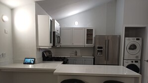 A modern kitchen with white cabinets, a stainless steel refrigerator, a microwave, and a built-in washing machine. There are two black stools at the kitchen island.