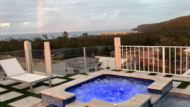 A swimming pool with a hot tub, surrounded by a deck with lounge chairs and a view of a distant cityscape and hills.