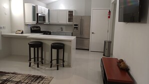 A modern kitchen with white cabinets, a stainless steel refrigerator, a microwave, and a gas stove. There are two black bar stools and a small rug on the floor.