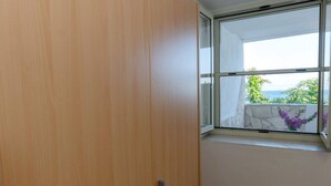 Een kamer met een houten garderobe, een klein wit krukje en een raam met uitzicht op groen en een gebouw.