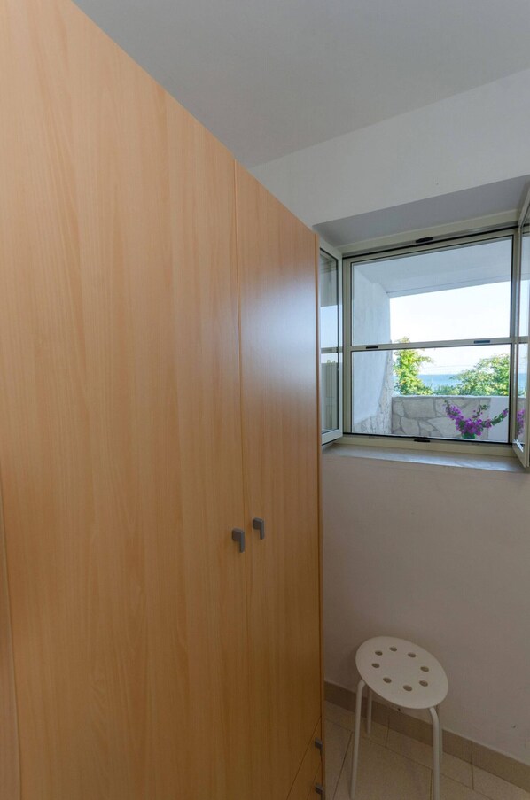 A room with a wooden wardrobe, a small white stool, and a window with a view of greenery and a building.