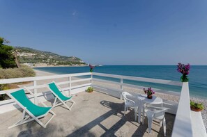 A balcony with white chairs and a table, overlooking a beach and clear blue sea.