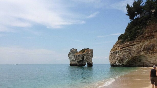 A beach with clear water, a large rock formation, and a person walking.