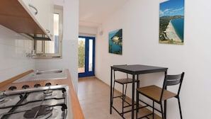 Una cocina con estufa, lavamanos y una puerta azul que da a un balcón. Hay una mesa con dos sillas y dos cuadros en la pared.
