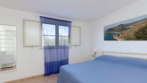 Un dormitorio con cama, una ventana con cortinas translúcidas, un cuadro de un paisaje costero y una mesita de noche blanca con lámpara.