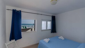 A bedroom with a bed, a window with curtains, a view of the sea, and a white chair. - Miramare House - Trilo 4 B (Mattinata)
