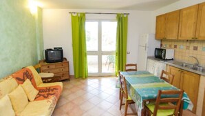 Ein kompakter Wohnraum mit einer Couch, einem Fernseher, einem Esstisch mit Stühlen, einer Küchenzeile inklusive Kühlschrank und einer Glastür zum Außenbereich.