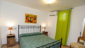 Ein Schlafzimmer mit einem Bett, zwei Nachttischen mit Lampen, einem Sessel, einem Bild an der Wand und einem Fenster mit grünem Vorhang.