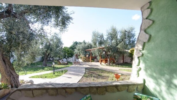 Eine Terrassenfläche mit einer Steinsitzbank, einem Tisch mit Blumenmuster-Tischdecke und Blick auf einen Garten mit Bäumen und einer Pergola.