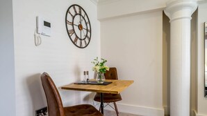 A dining area with a wooden table, two brown leather chairs, a vase with flowers, a wall-mounted clock, and a white column.