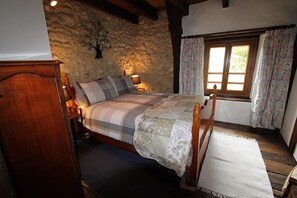 A cozy bedroom with a wooden bed, a stone wall, a window with floral curtains, and a nightstand with a lamp.