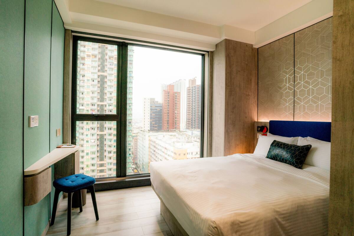 A modern hotel room with a large window, a bed with a geometric-patterned headboard, and a small desk with a chair.